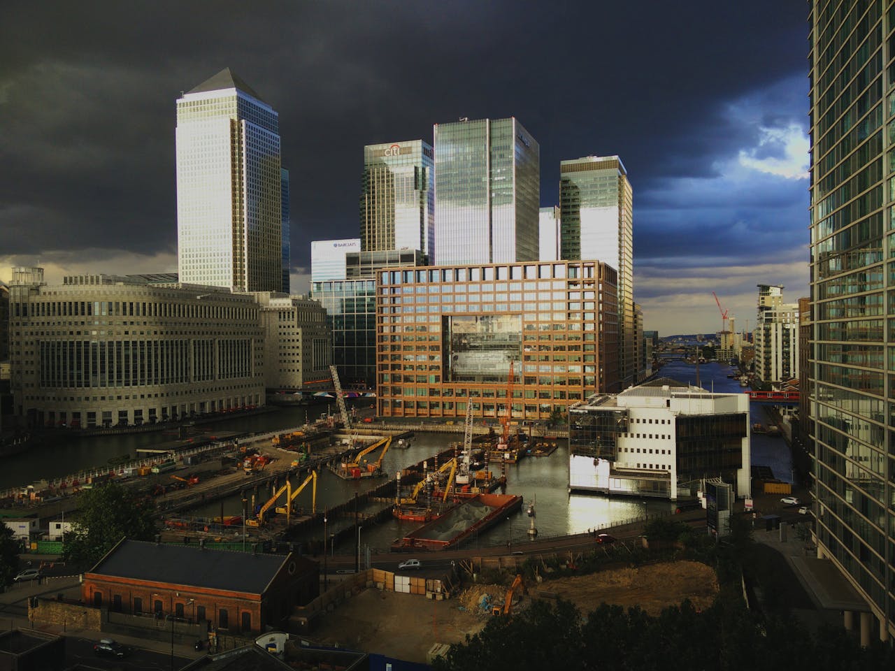 A dramatic scene of Canary Wharf skyscrapers and construction site against a dark, stormy sky.